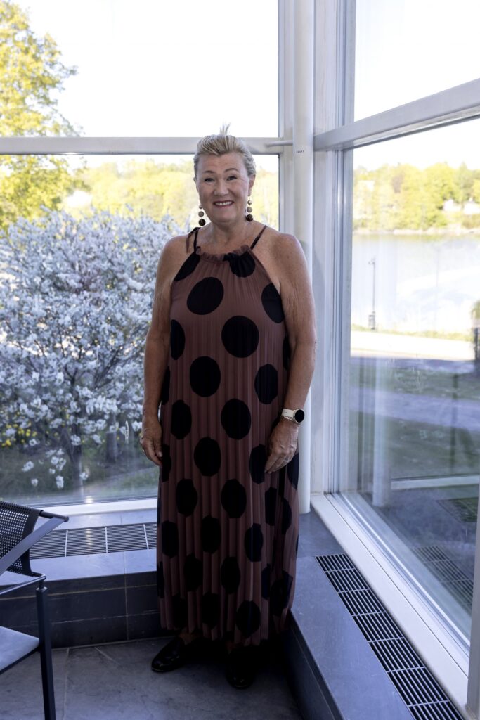 Vaaleahiuksinen henkilö hymyilee Oopperatalon päälämpiön ikkunoiden edessä. Hänellä on yllään nilkkoihin asti ulottuva ruskea kesämekko, jossa on mustia palloja.