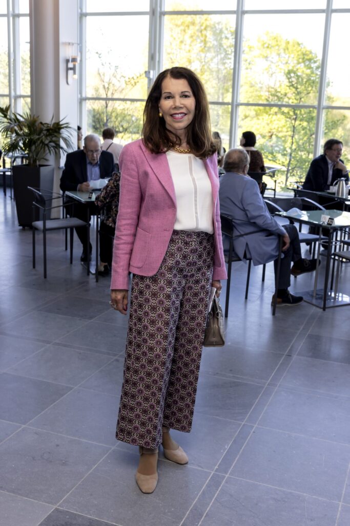 Henkilö hymyilee ja poseeraa kameralle Oopperatalon päälämpiössä. Hänellä on hartioille ulottuvat ruskeat hiukset, vaaleanpunainen jakku ja kuviolliset housut.