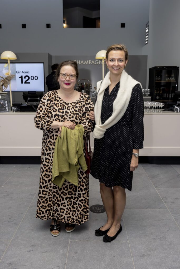 Kaksi hymyilevää henkilöä seisoo vierekkäin Oopperatalon samppanjabaarin edustalla. Toisella heistä on suuret silmälasit, leopardikuvioinen mekko ja vihreä villatakki, toisella on yllään musta pilkullinen mekko ja valkoinen villatakki kiedottuna harteille.